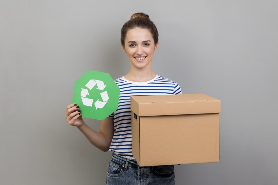 A woman stands holding a green recycling logo in one hand and a brown corrugated box in the other.